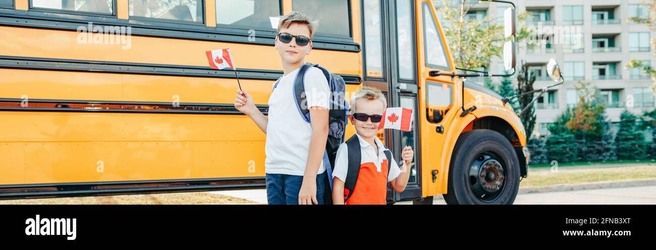 Back to school. Happy Caucasian boys students holding waving Canadian flag. Students kids by yellow school bus outdoors. Education and back to school Stock Photo