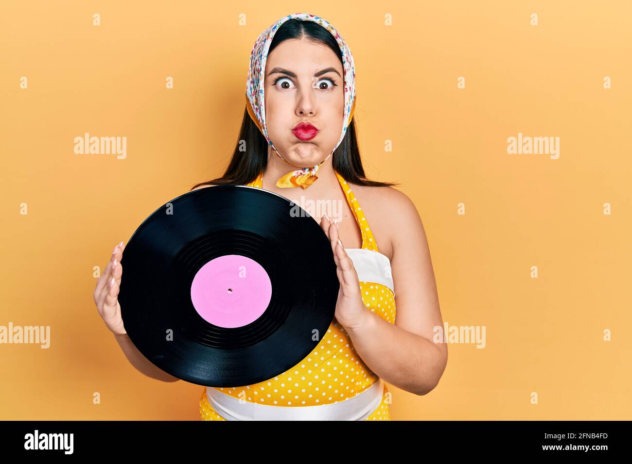 Young hispanic woman wearing pin up style holding vinyl disc puffing cheeks with funny face. mouth inflated with air, catching air. Stock Photo