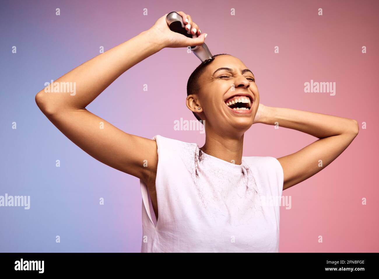 Smiling woman shaving her head. Bold and liberated female cutting her hair with an electric trimmer against multicolored background. Stock Photo