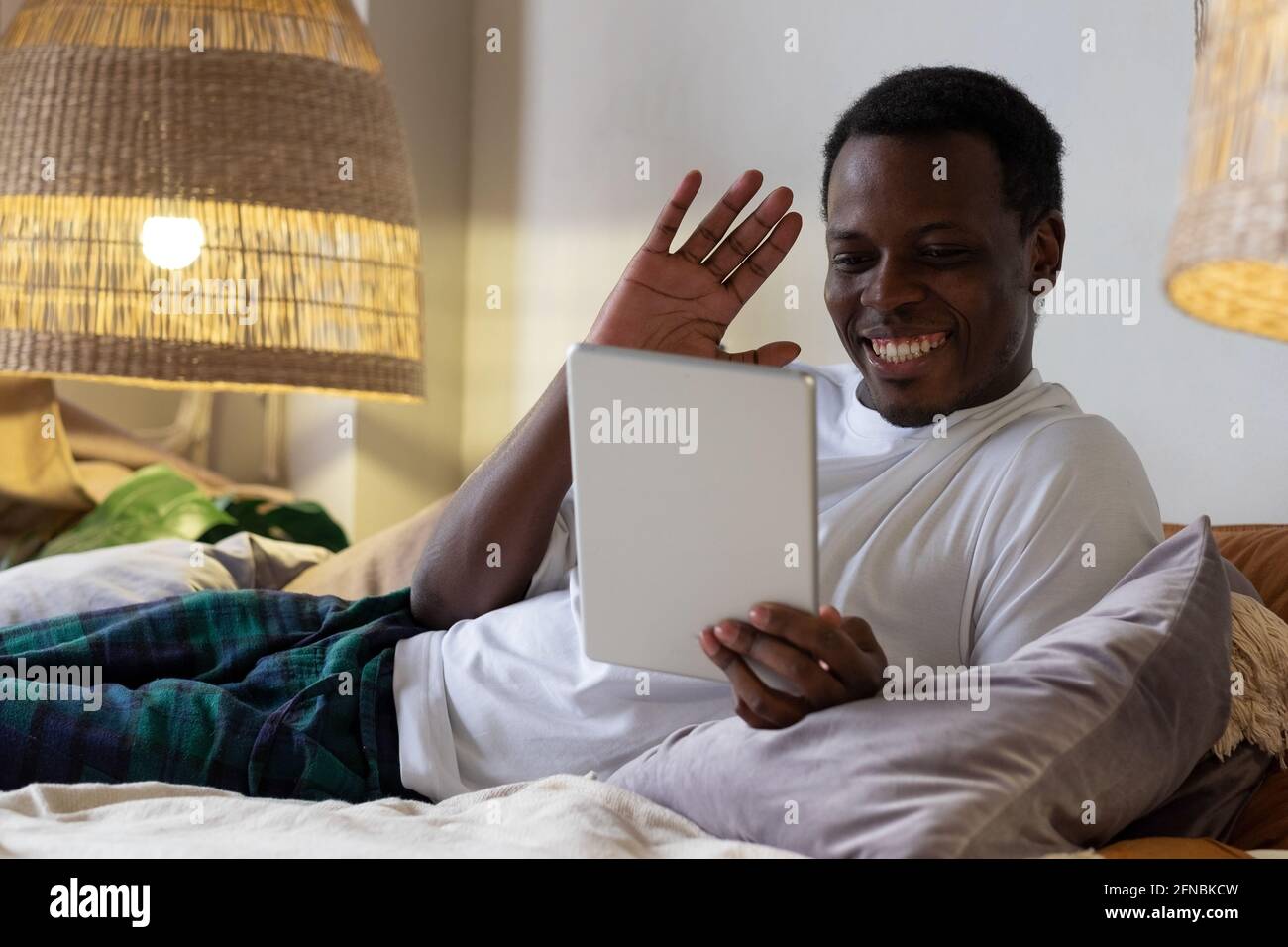 Black man with tablet resting in bed greeting waving hand. Stock Photo