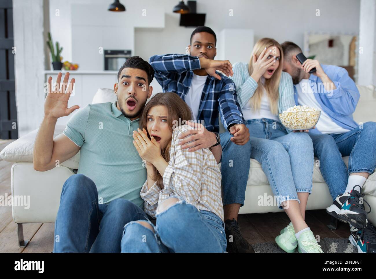 Frightened millennial friends watching scary movie or thriller, holding popcorn, covering their faces in horror at home Stock Photo