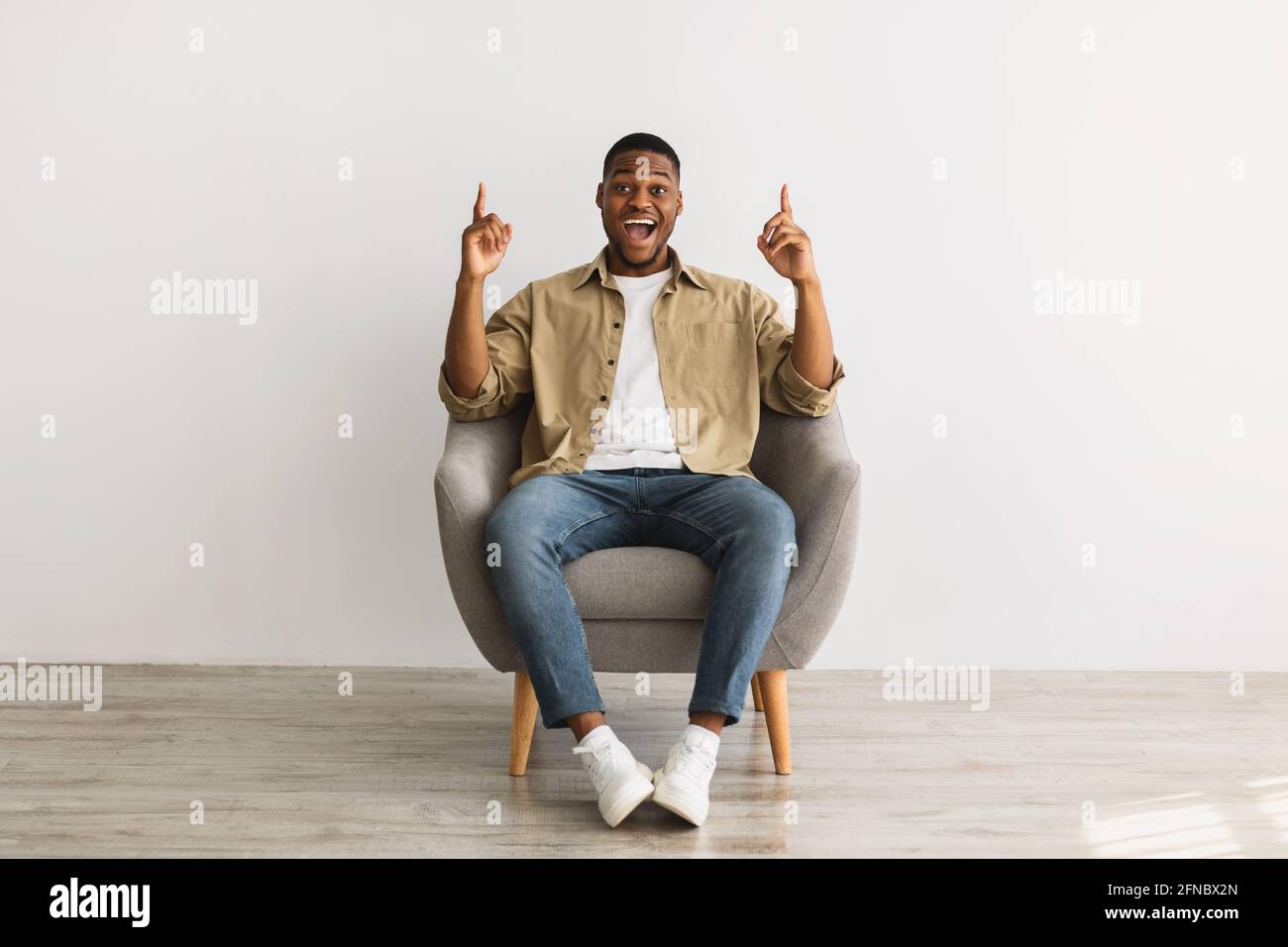 Excited African Millennial Man Pointing Fingers Up Sitting, Gray Background Stock Photo