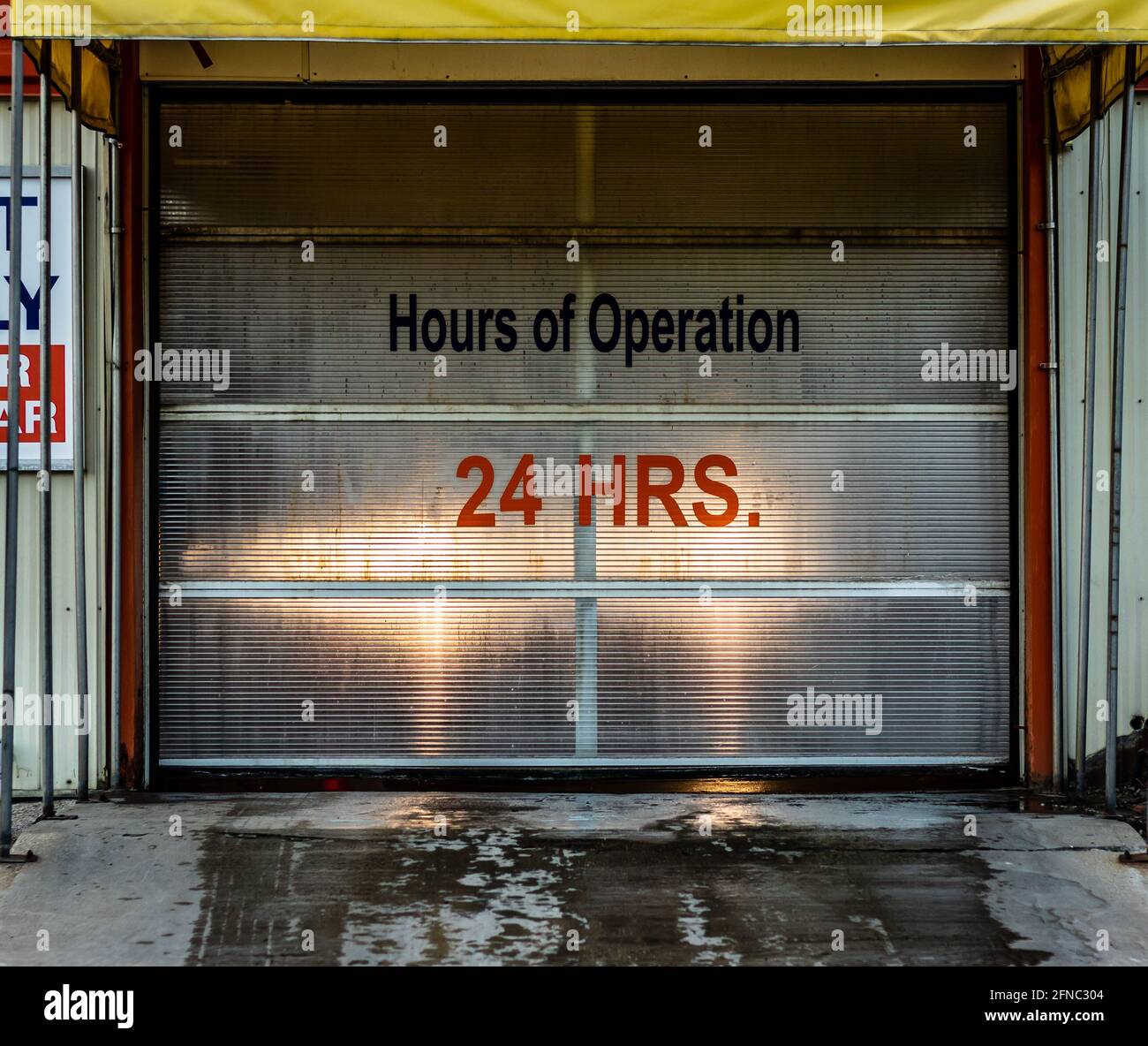 Hours of operation (24 hours) vinyl letter and number sign on roll door of car wash. Headlights shining through, tire marks on pavement. Stock Photo
