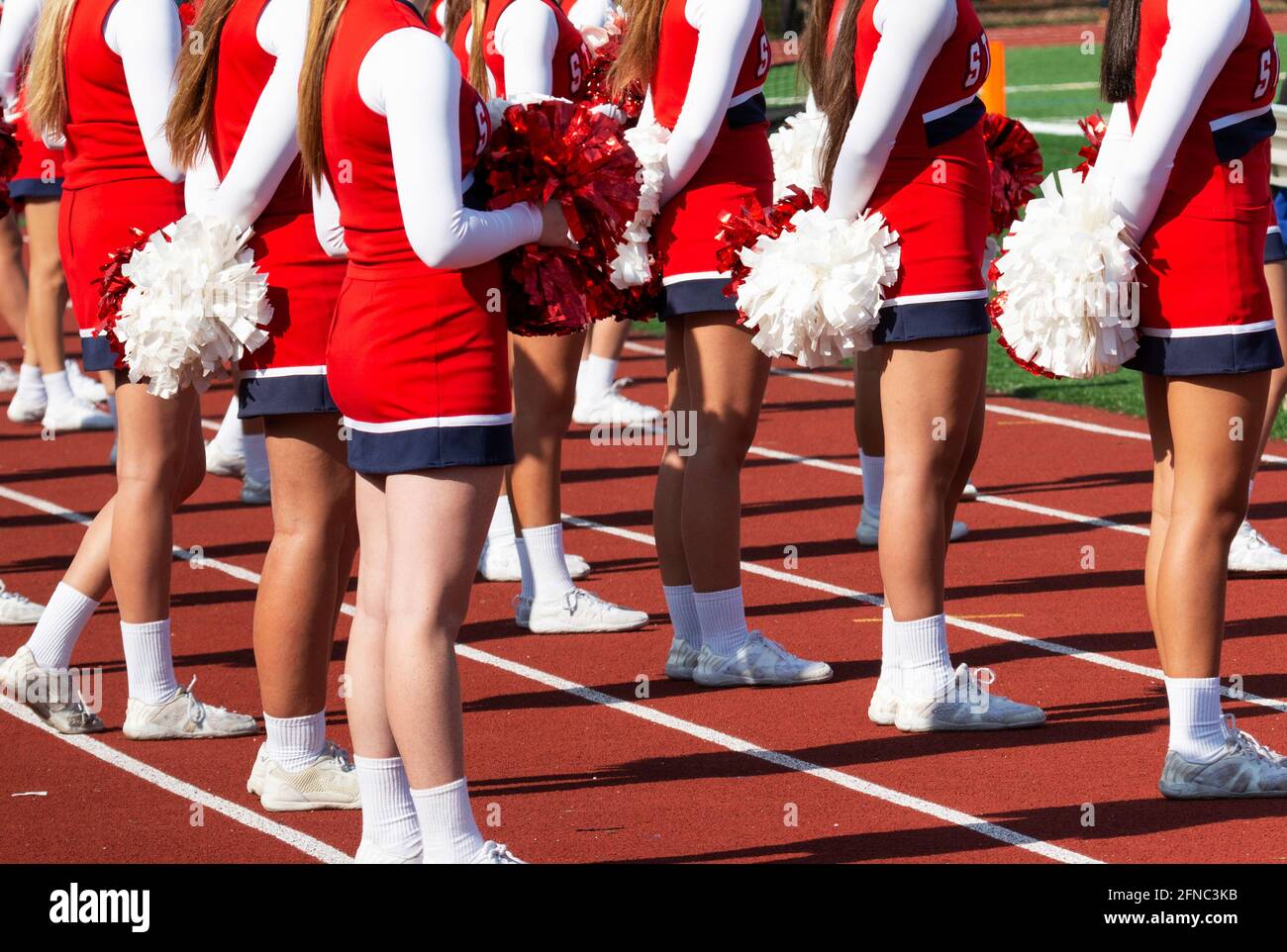 Rear view of high school cheerleaders holding pom poms standing on a red track facing the field during a football game. Stock Photo