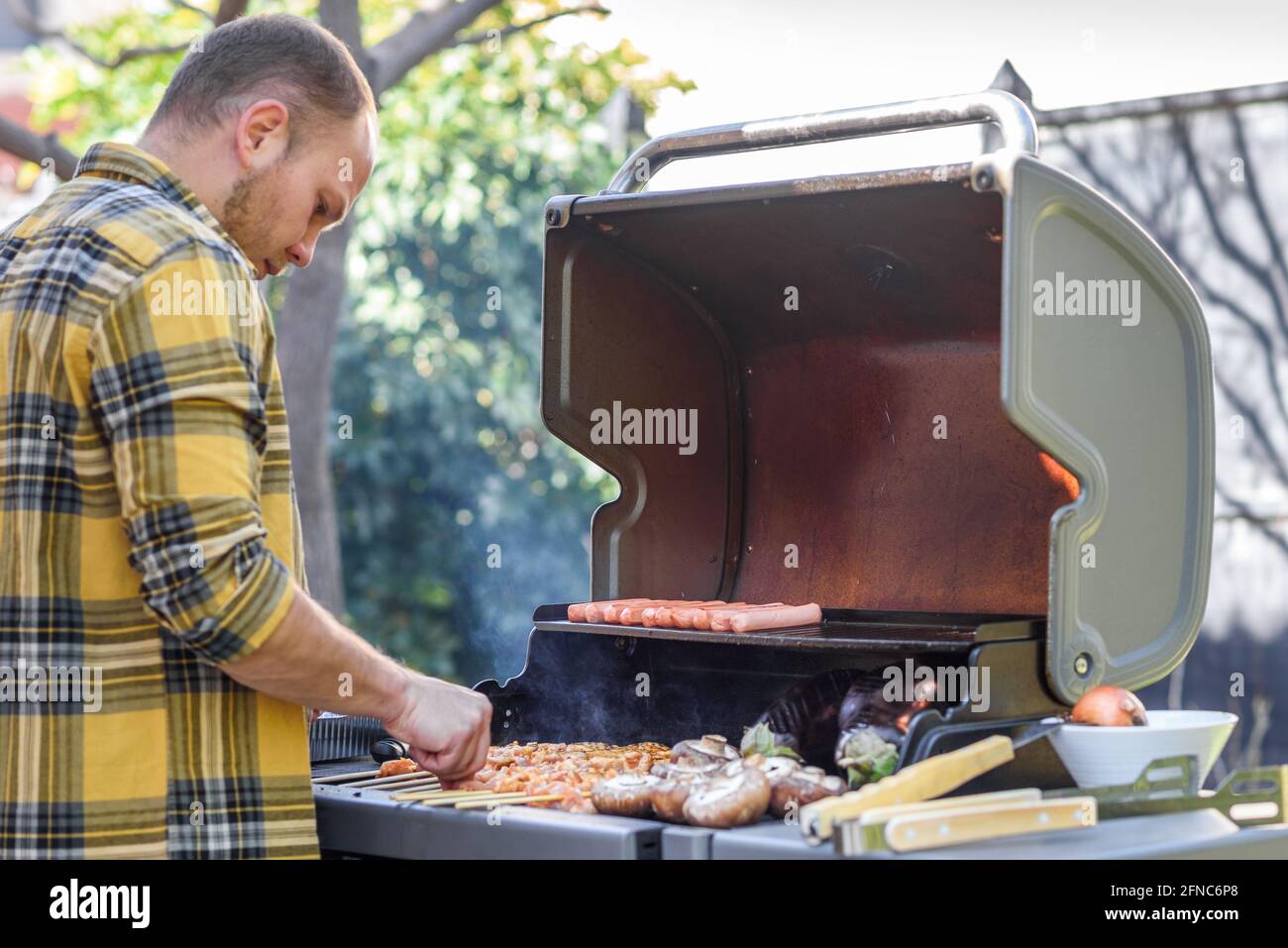 Man outside with bbq. Young man grilling meat skewers, meat steak, eggplant, and mushrooms on a gas grill. Selective focus is on sausage and steak. Stock Photo