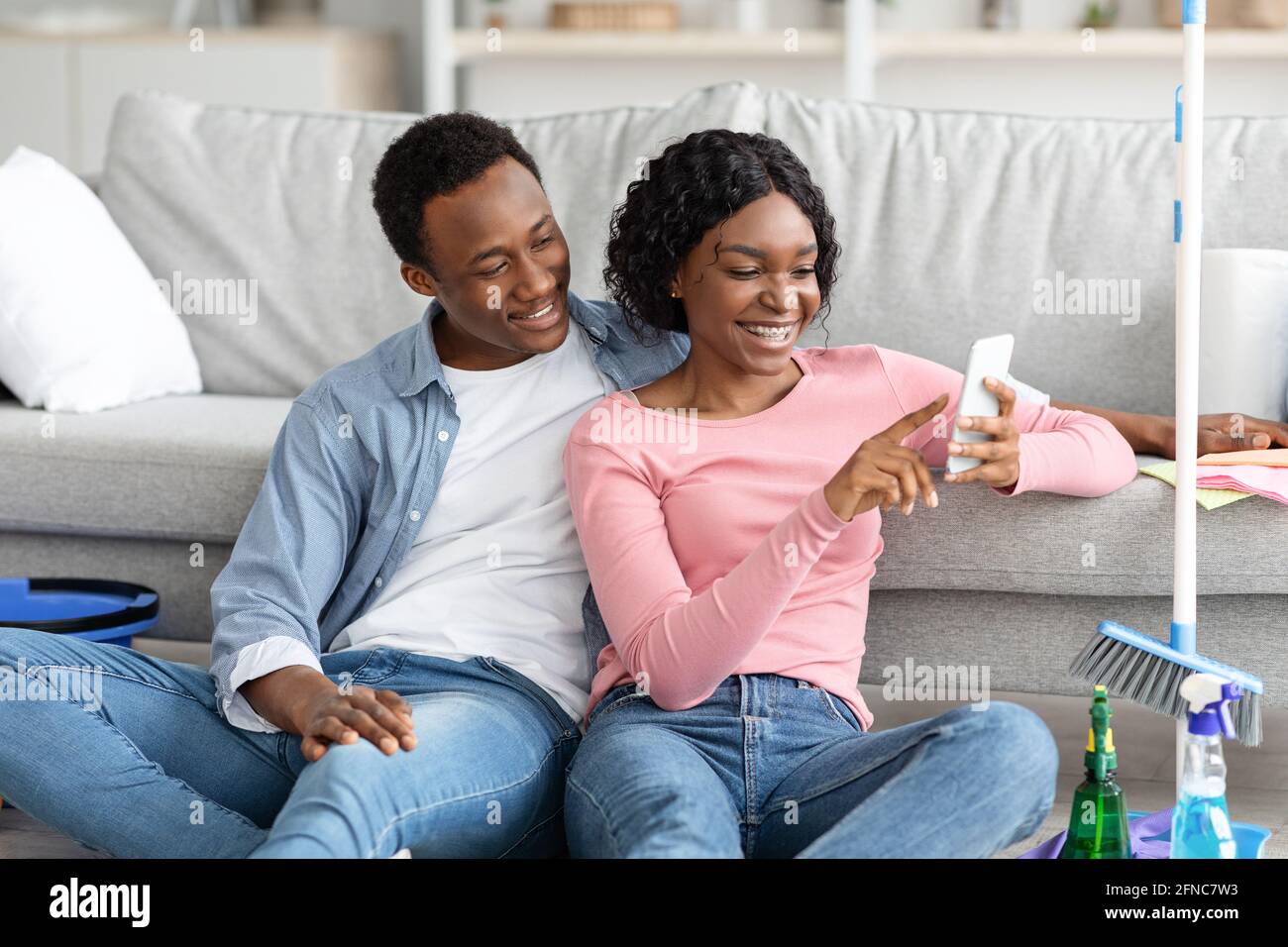 Black lovers with cleaning tools sitting on floor, using smartphone Stock Photo