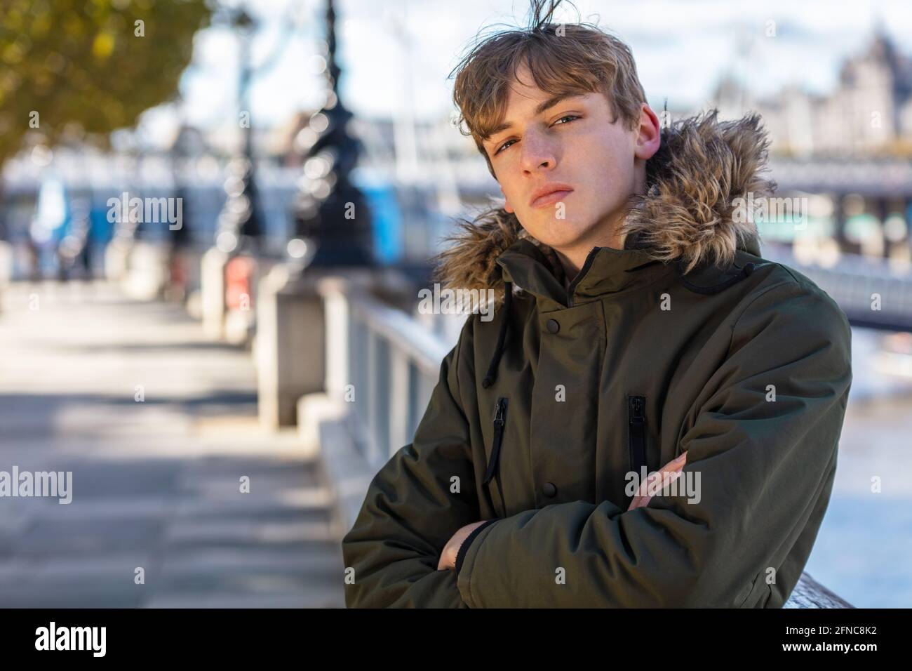 Young adult male boy teenager outside in sunshine wearing a hooded parka coat in an urban city Stock Photo