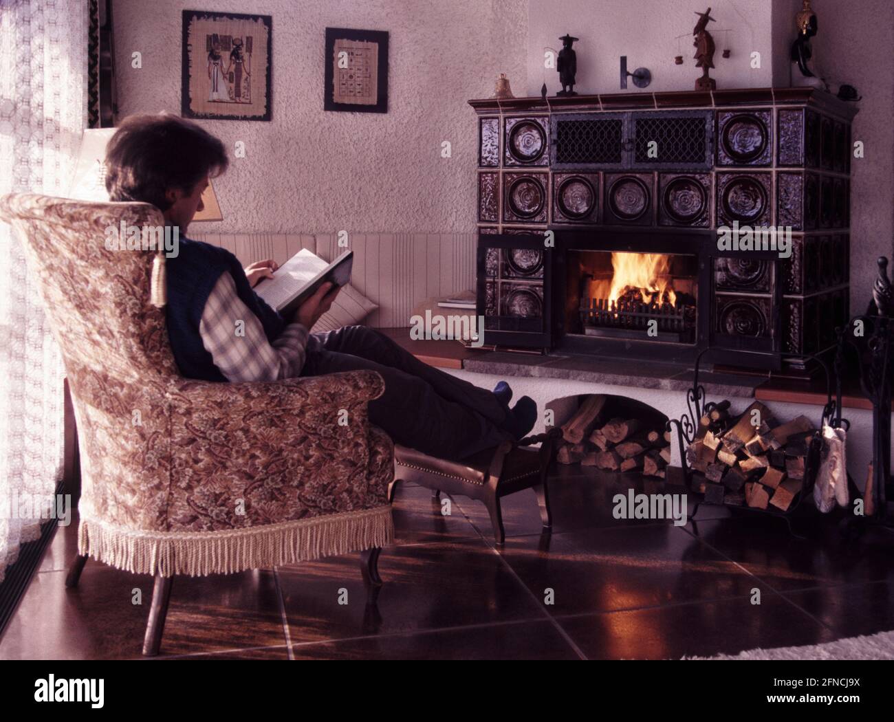ein Mann liest ein Buch in einem alten Lehnsessel am Kaminfeuer, Sonne scheint mild von links * man relaxed reading a book in front of fireplace Stock Photo