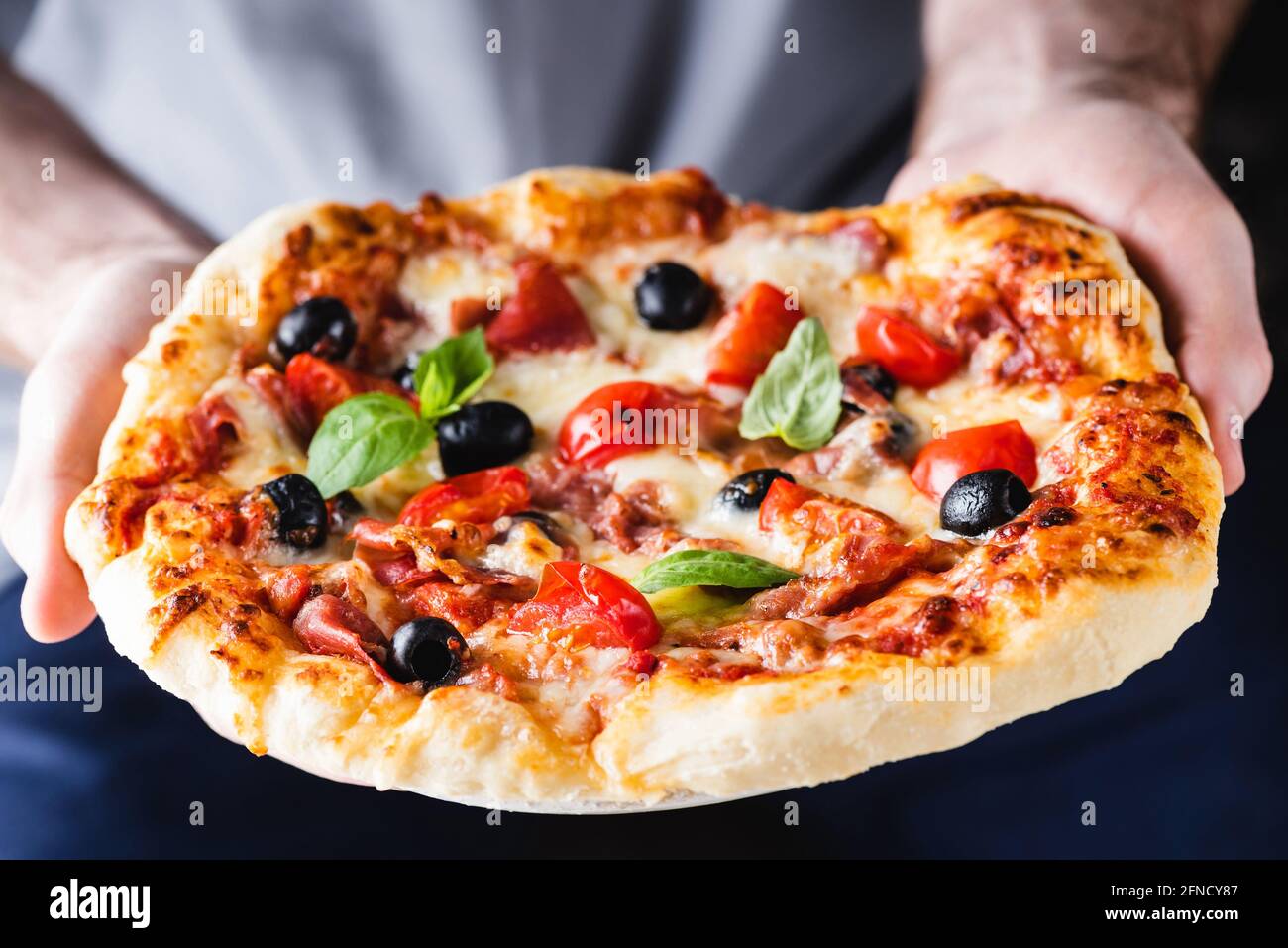 Male hands holding a pizza. Baked hot italian pizza with ham, black olives and tomatoes in hands Stock Photo