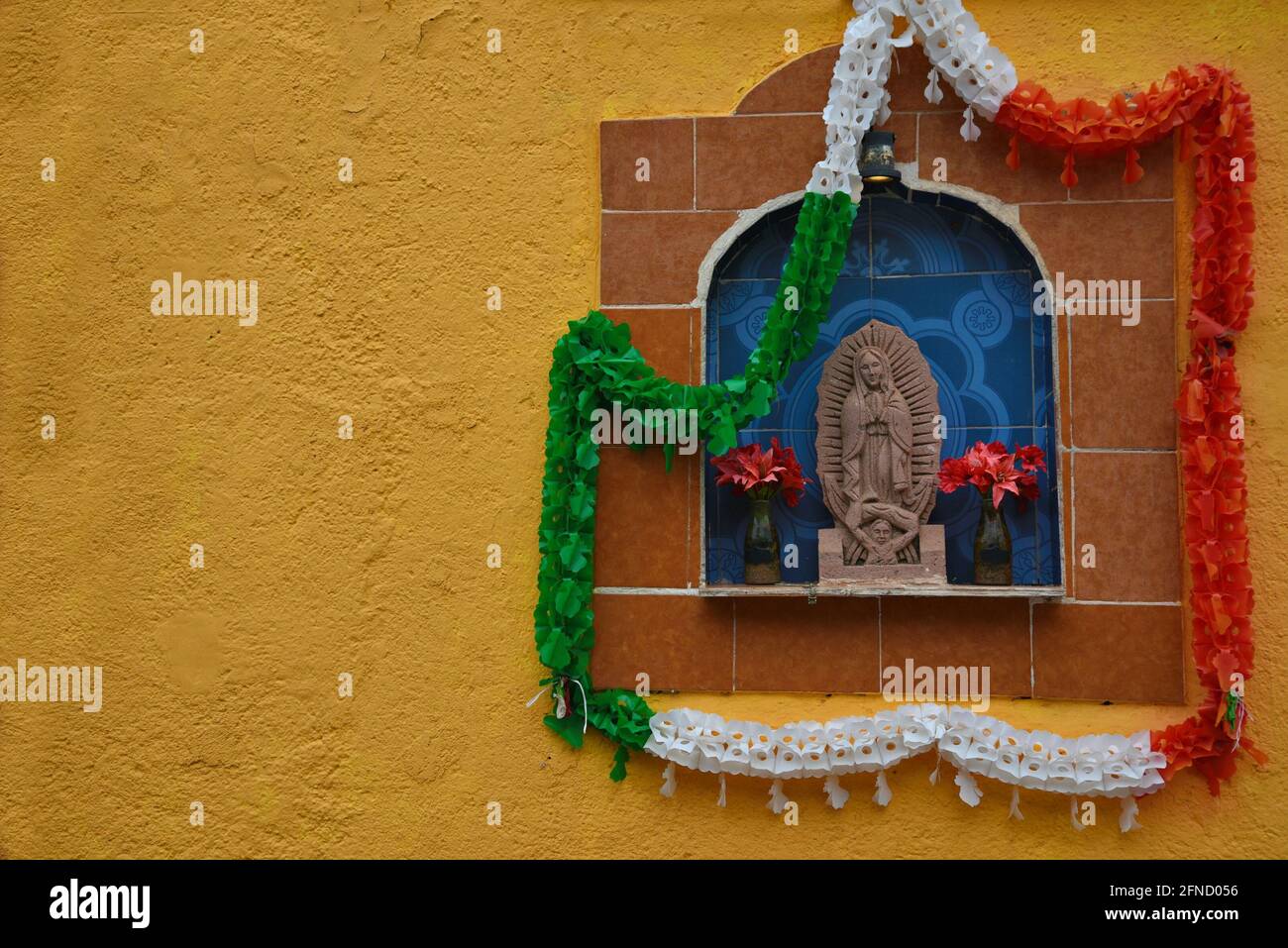 Ceramic tile religious icon decorated with a Mexican flag colors garland on an ochre stucco wall in the ghost town of Real de Catorce, Mexico. Stock Photo