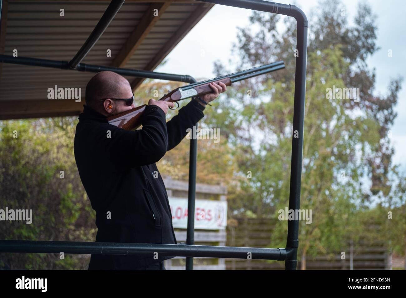 Bald man wearing safety sunglasses and earbuds holding a shotgun