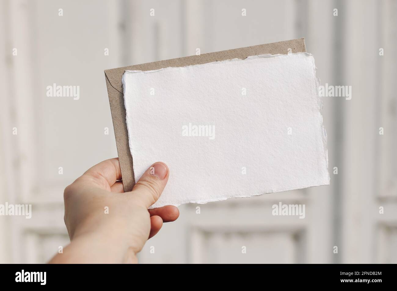 Bright feminine stationery mockup scene. Closeup of woman hand holding handmade paper greeting card, envelope. Blurred white old wooden doors Stock Photo