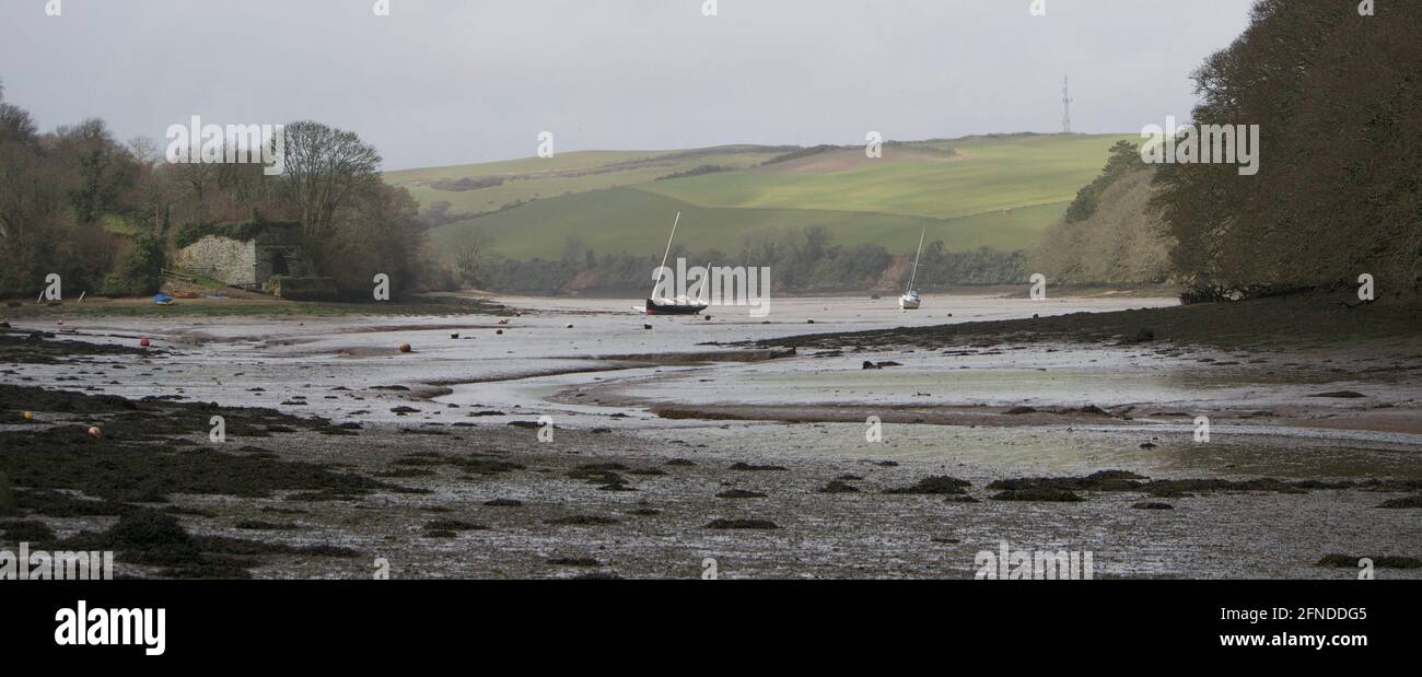 Muddy, tidal estuary at low tide. Green fields and rolling hills in the misty distance; seaweed in the foreground and sailing boats moored on buoys (a Stock Photo