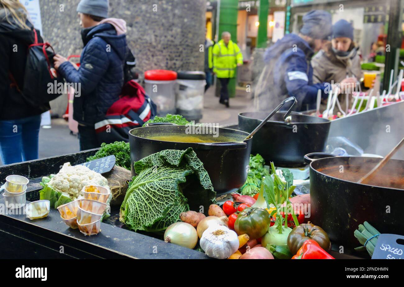 London UK 1 10 2018 Vegetables piled up around cast iron cooking pots full of steaming soup as customers stand around at famous Boroughs Market Stock Photo