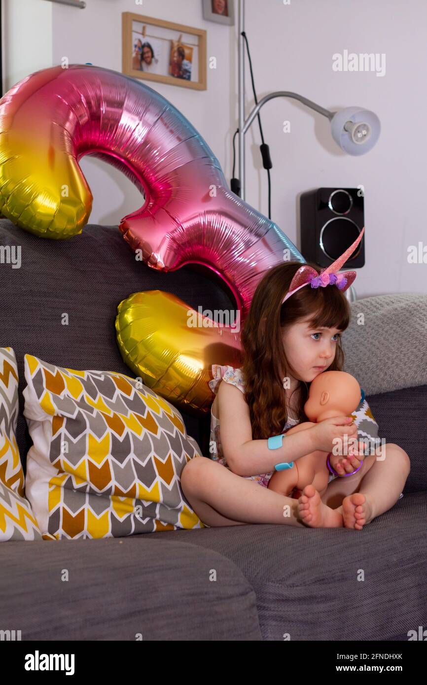 A Cute Baby Girl Sitting On A Sofa With A Colourful Balloon And A Doll Stock Photo