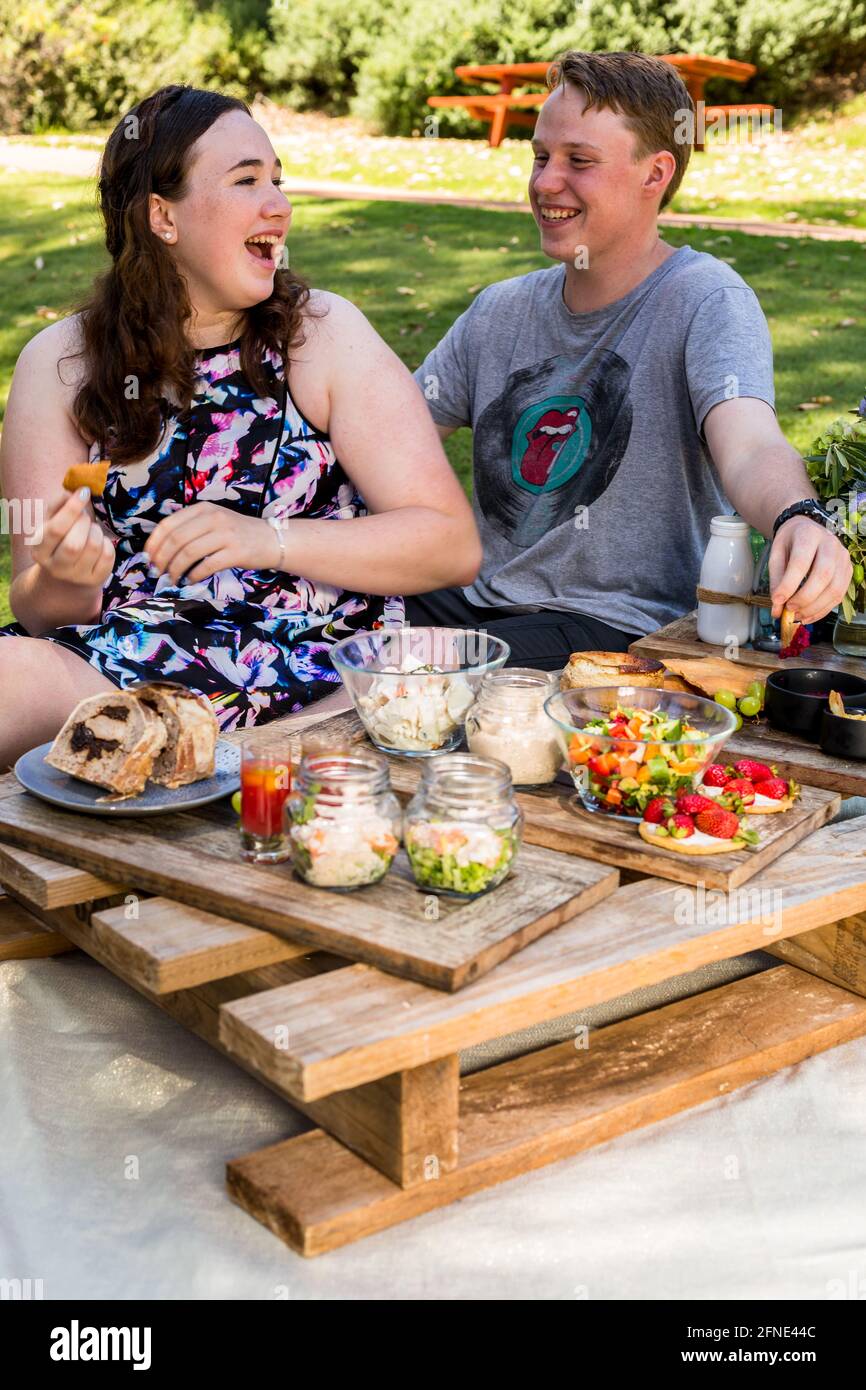 young couple laughing while sitting on grass in park having gourmet picnic Stock Photo