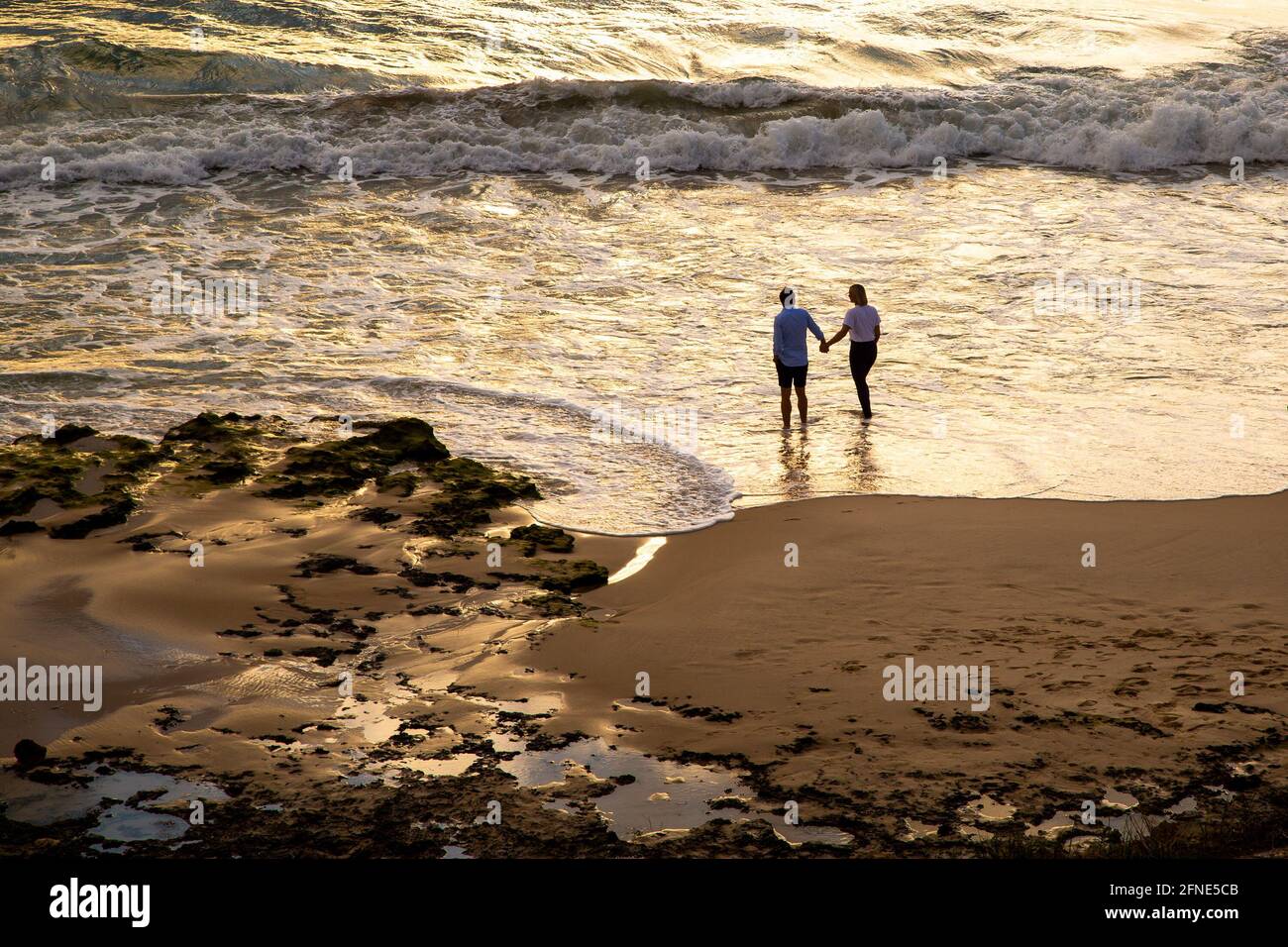 Couple holding hands on beach with golden light reflecting on ocean and sand. People facing away from camera looking at sunset Stock Photo