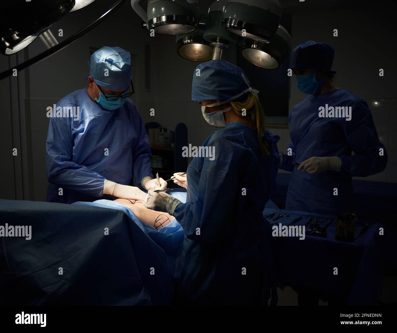 Medical team doing aesthetic plastic surgery in operating room with dim light. Surgeon and assistants performing cosmetic surgery and using medical instruments. Concept of medicine and plastic surgery Stock Photo