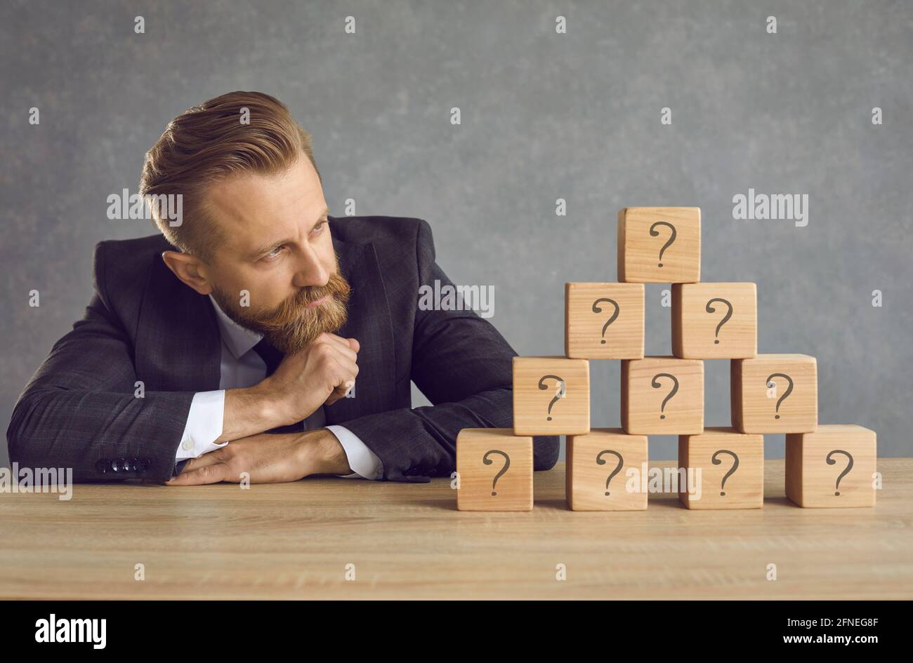 Businessman in suit looking at pyramid of cubes with question marks and thinking of answer Stock Photo