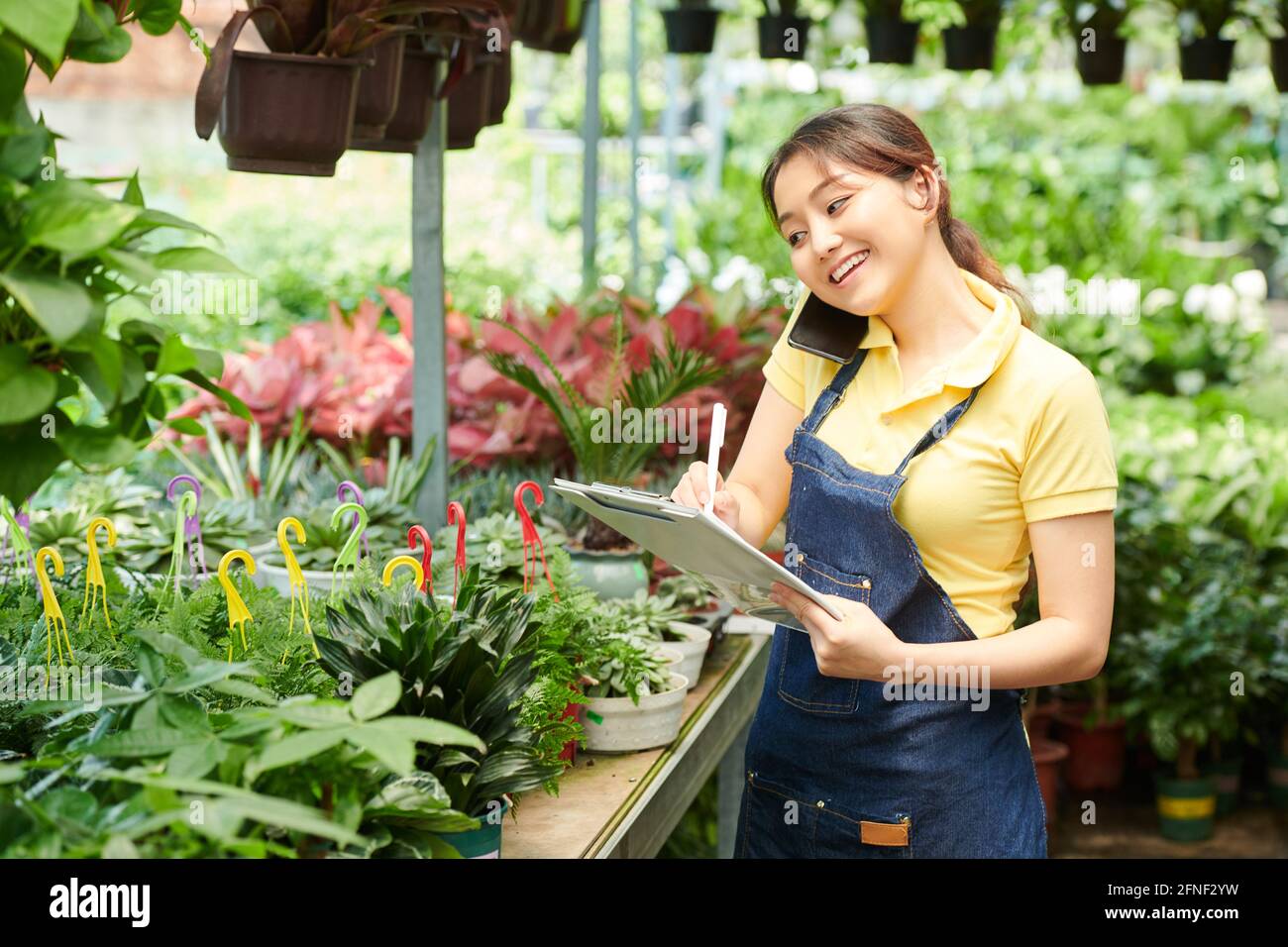Smiling pretty gardening center worker talking on phone with customer and taking notes in document Stock Photo