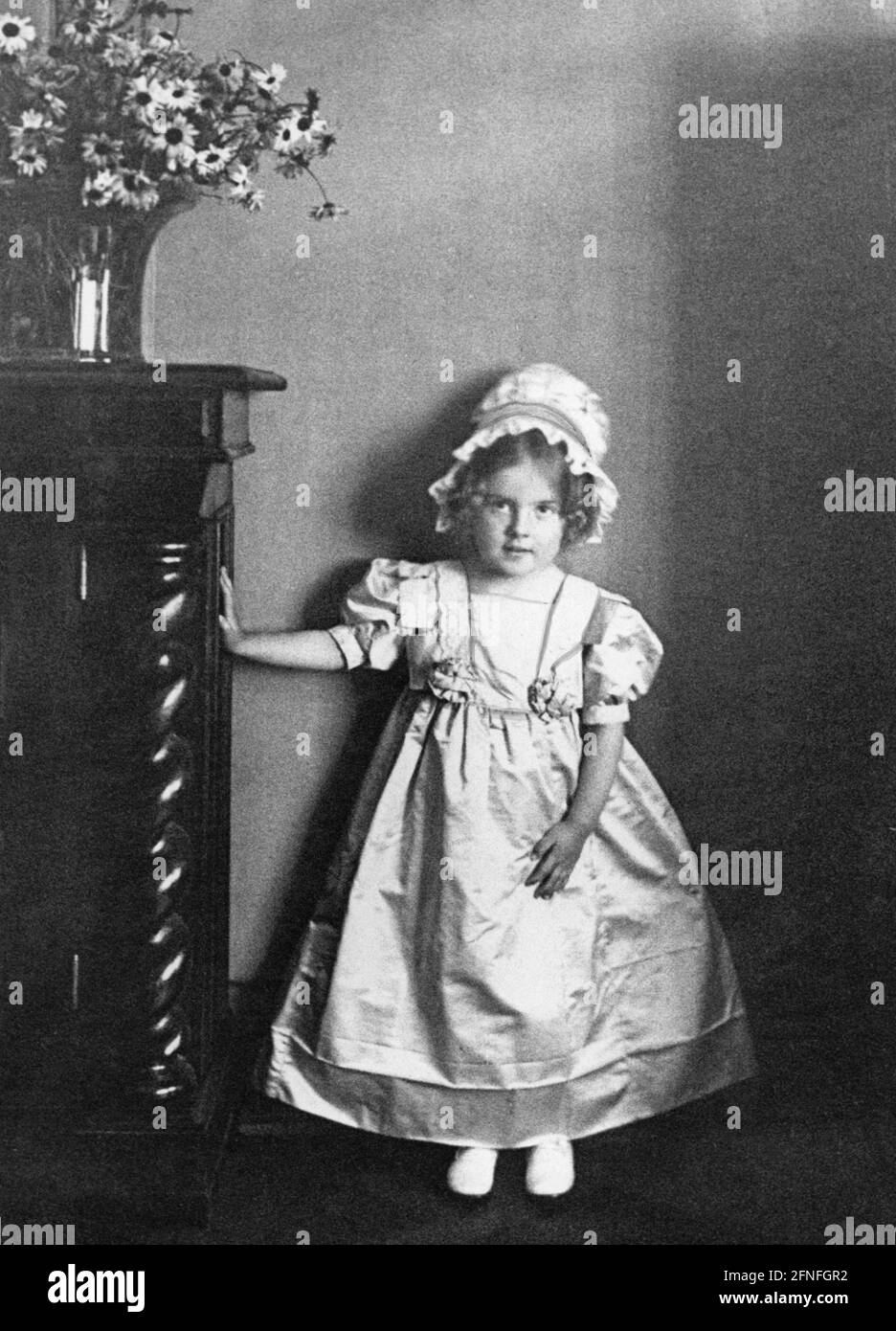 A little girl in a dress leans with outstretched arm against a cupboard on which a vase with flowers stands. Undated photograph, ca. 1900. [automated translation] Stock Photo