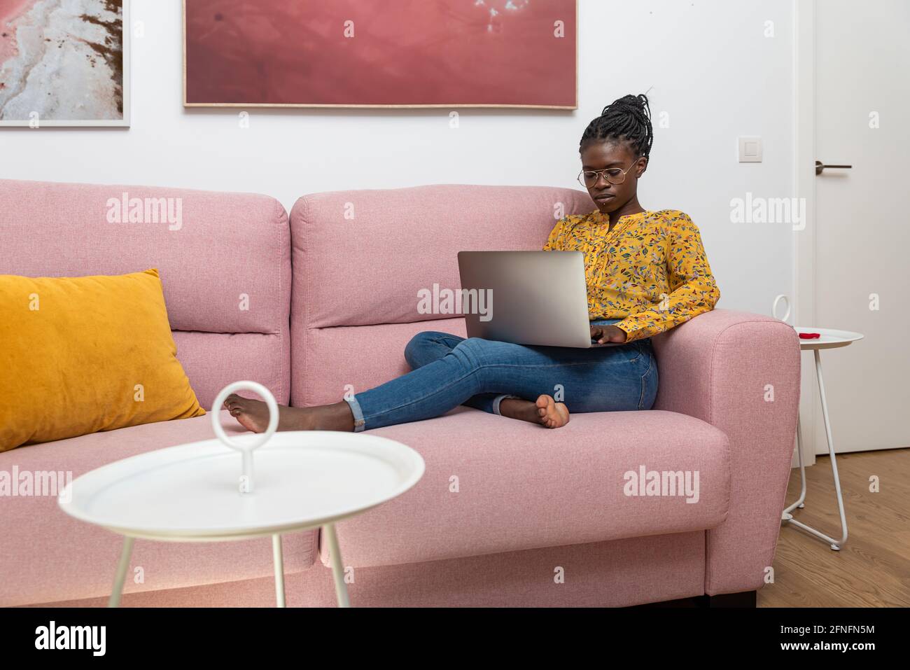 Focused African American female freelancer sitting on sofa at home and typing on netbook while working on project Stock Photo