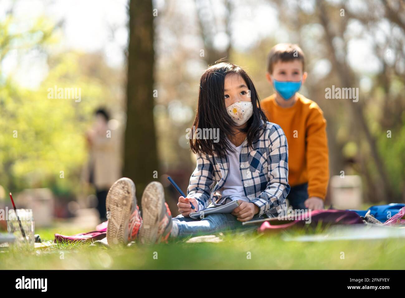 Small children sitting outdoors in city park, learning group education and coronavirus concept. Stock Photo