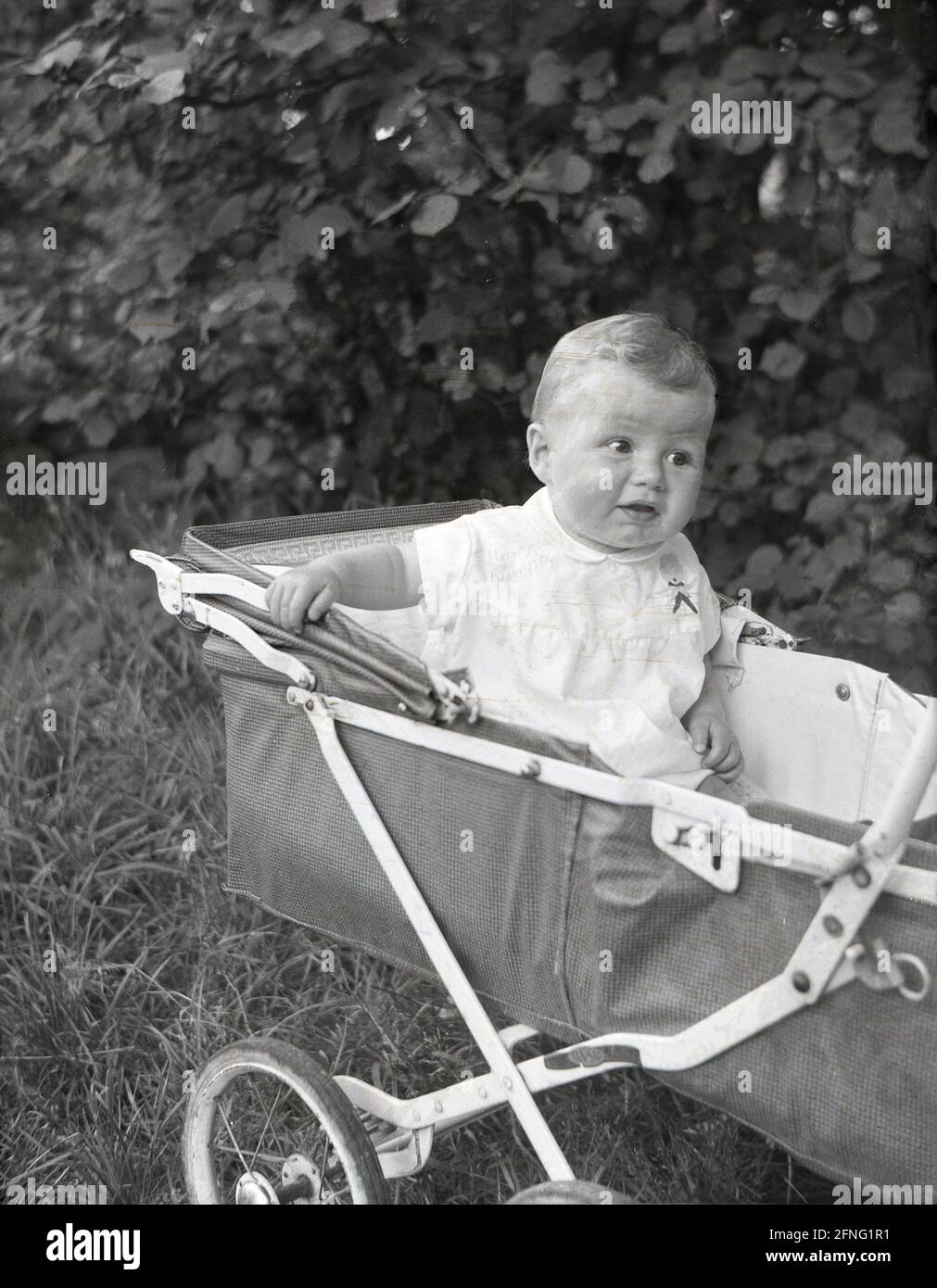 1950s, historical, outside parked on some grass, an infant boy sitting up in a canvas covered, metal framed, wire-wheeled, folding pushchair of the era, made by Tan-Sad, England, UK. Founded in Birmingham in 1922, Tan-Sad made constructional toys, metal factory chairs and childrens push chairs. In the 1950s and 60s the Tan-Sad name became so well-known, that it became a generic name for pushchairs. The company folded in 1975. Stock Photo