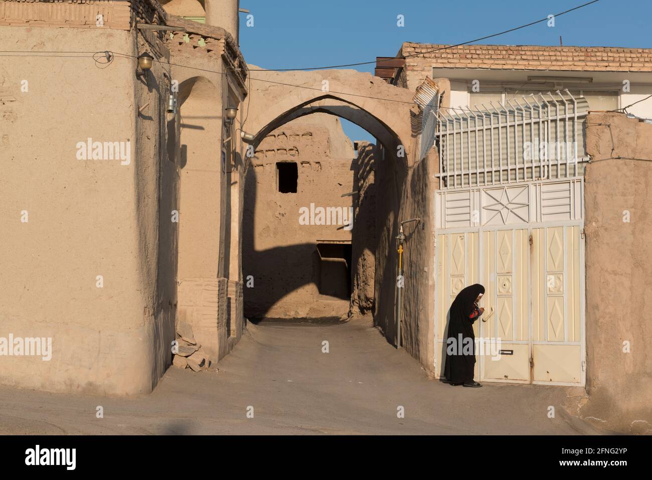 Senior woman in black chador opening a metal door in the late afternoon sun in the village Mohammadieh near Nain, Nain County, Isfahan Province, Iran Stock Photo