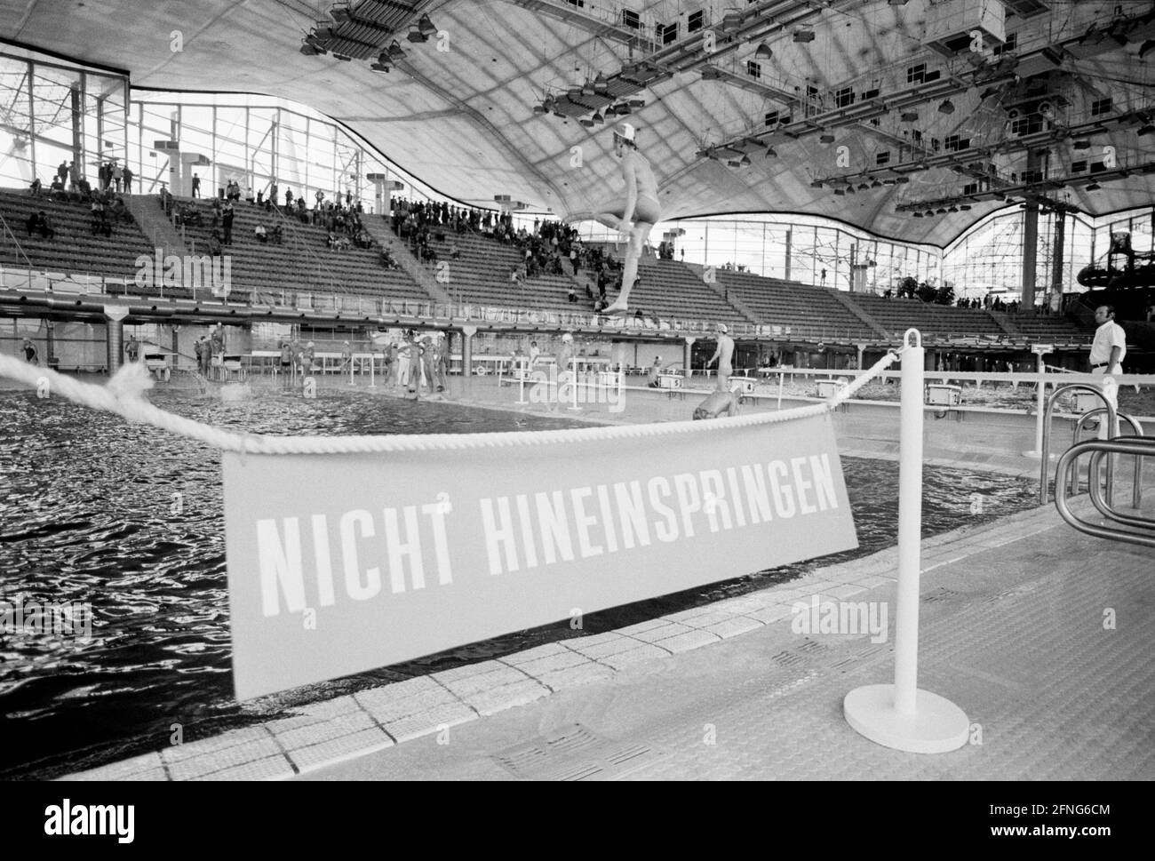 'At the edge of the pool hangs a sign on a string with the inscription ''Do not jump in''. In the background you can see a boy jumping into the pool from a diving tower. [automated translation]' Stock Photo