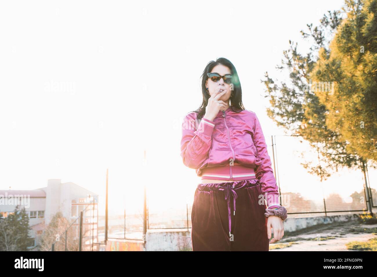 Millennial female in trendy wear and sunglasses smoking cigarette against urban building under white sky Stock Photo