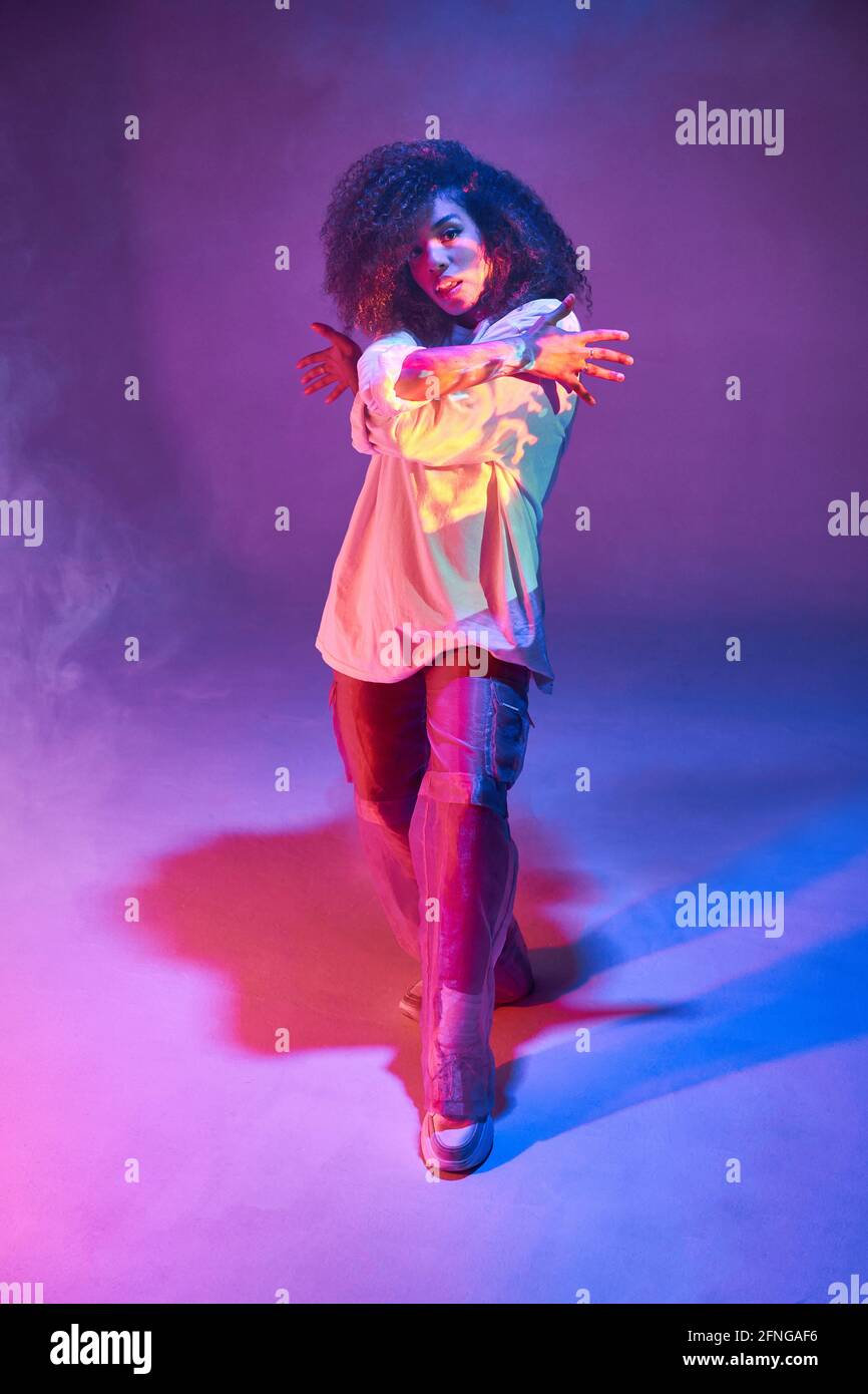 Full length fit young African American female dancer in loose informal wear  looking at camera while dancing in dark studio in neon lights Stock Photo -  Alamy