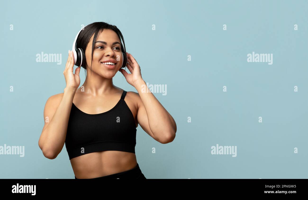 Active free time. Sporty black lady in sports uniform with headphones exercising or dancing, blue background, free space Stock Photo