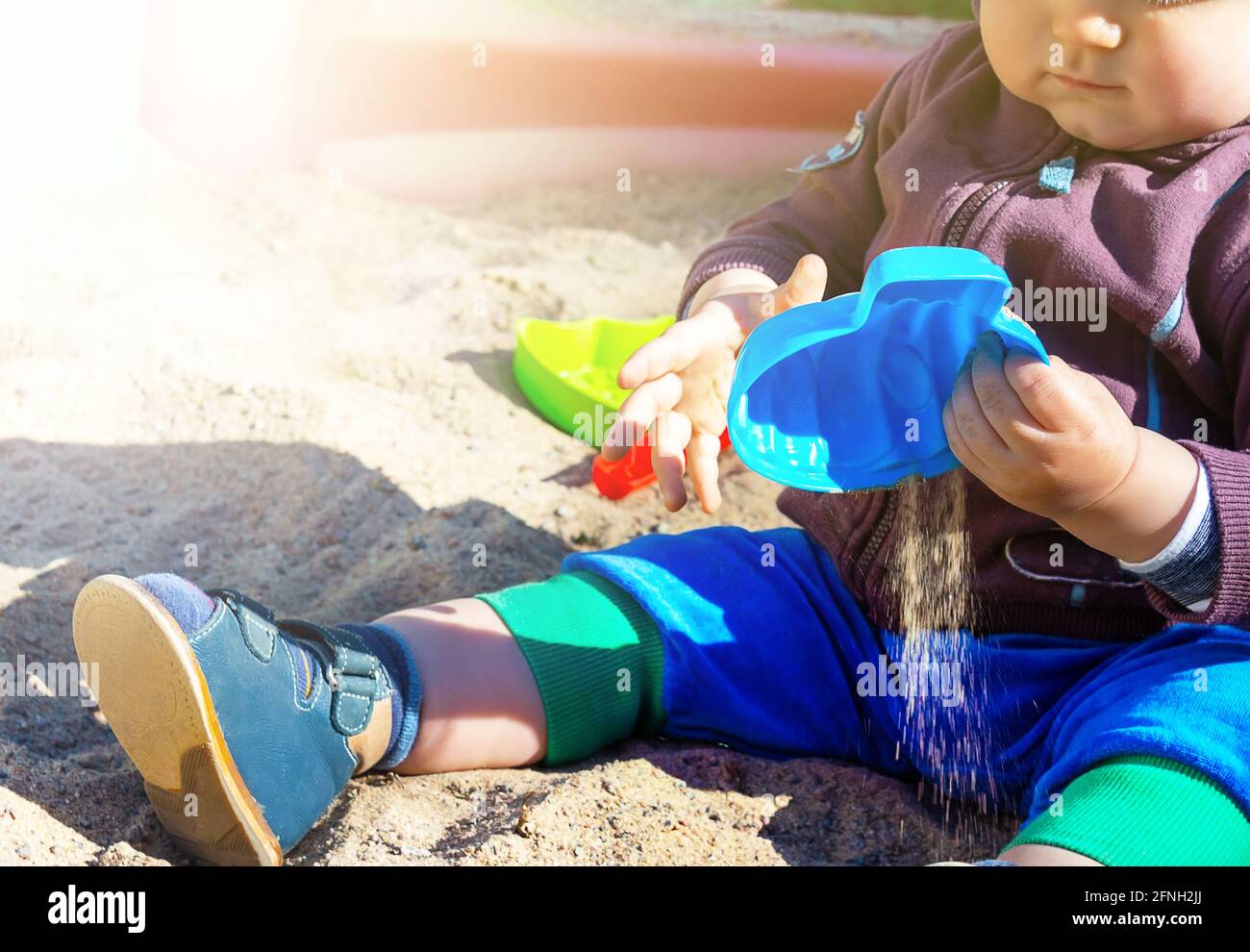 Unrecognizable little caucasian boy sitting in sandbox with toys on sunny summer day in big city. Child holding a blue sand mold.  Stock Photo