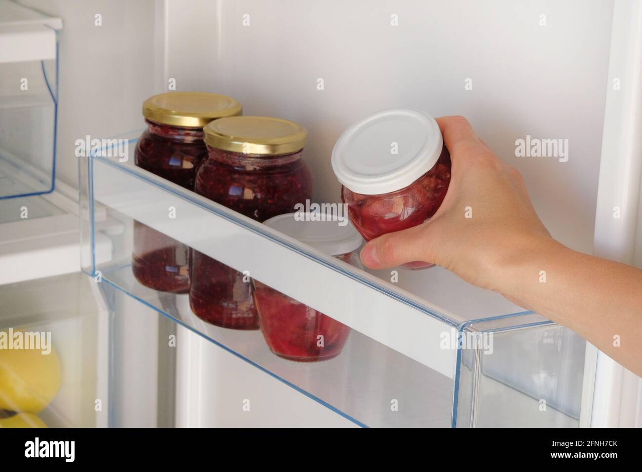 Glass Jars with raspberry and apple Jam on shelf in fridge. Female hand with a jar of red homemade jam. Fermented healthy foods stand in refrigerator. Stock Photo