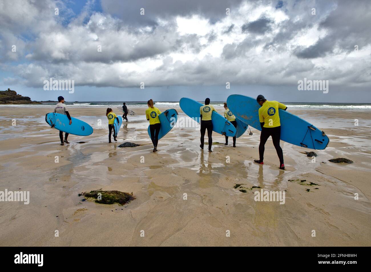 surfers carrying blue boards and wearing yellow rash vests and wet suits walking towards the sea along a flat expanse of wet sand on a cloudy morning Stock Photo