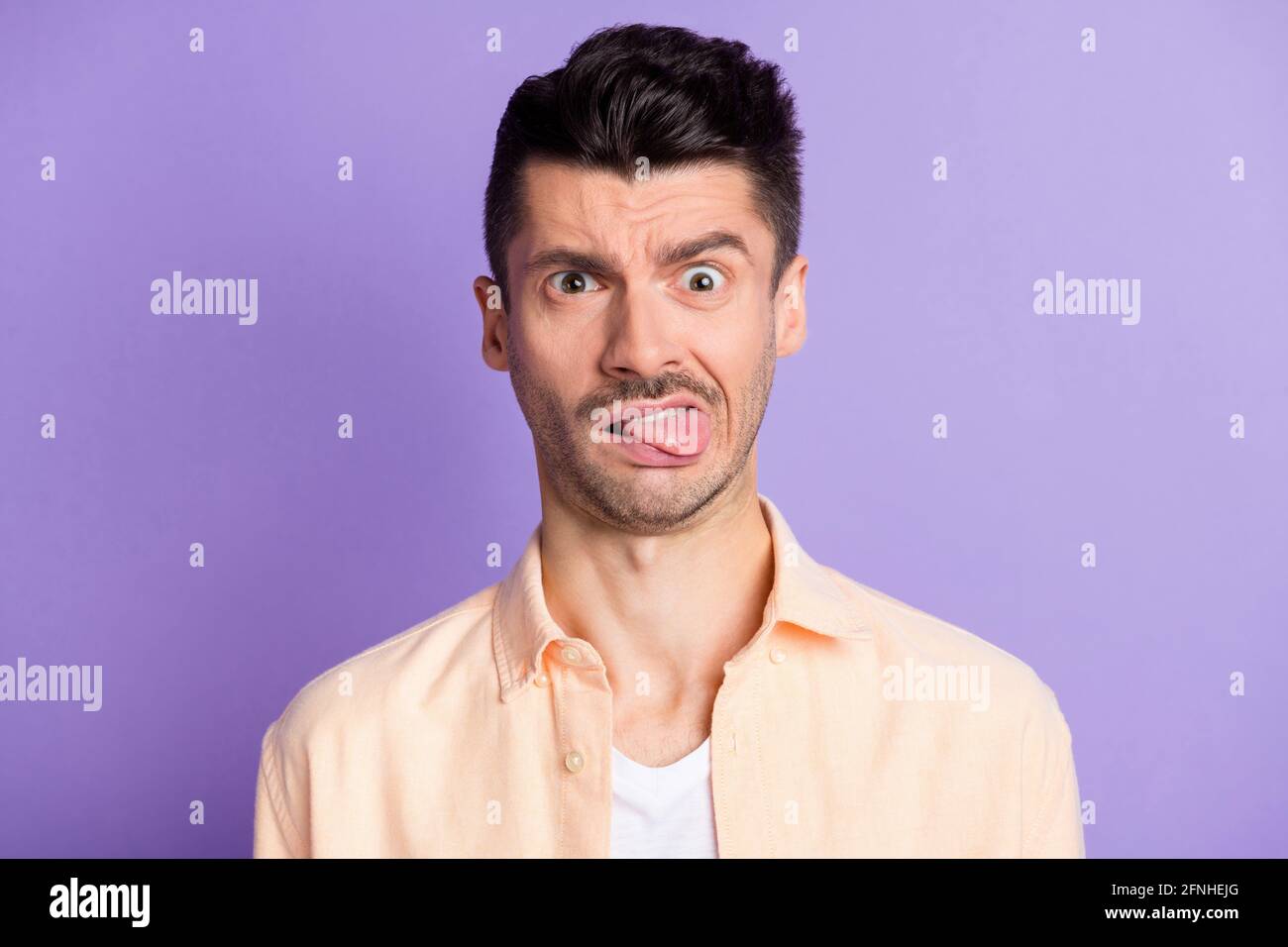 Photo of suspicious disgusted guy stick tongue out wear beige shirt isolated purple color background Stock Photo