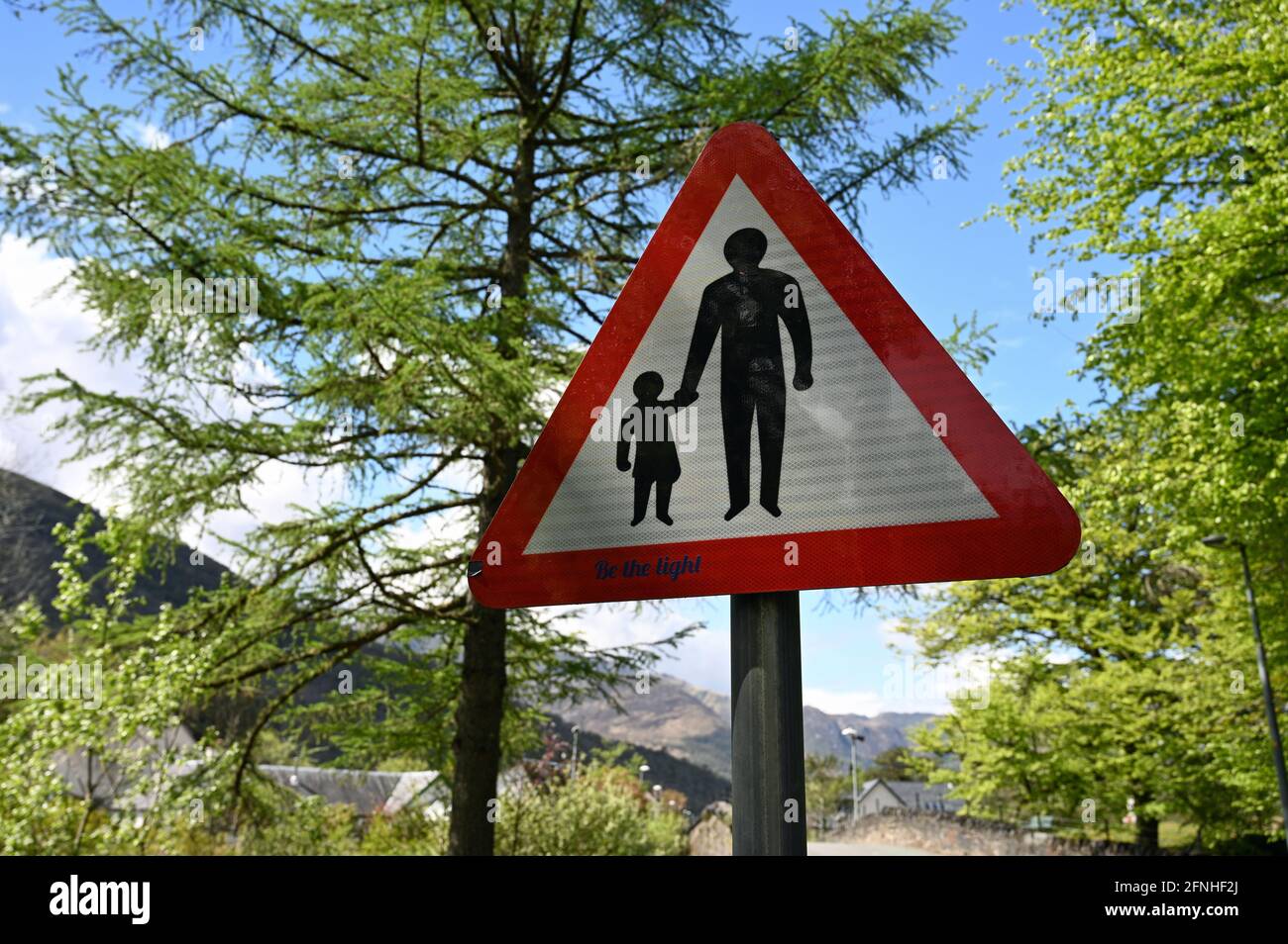 UK traffic sign for pedestrians with person holding hand of child. Blurred background of trees, mountains and quiet village road. Sunny day Stock Photo