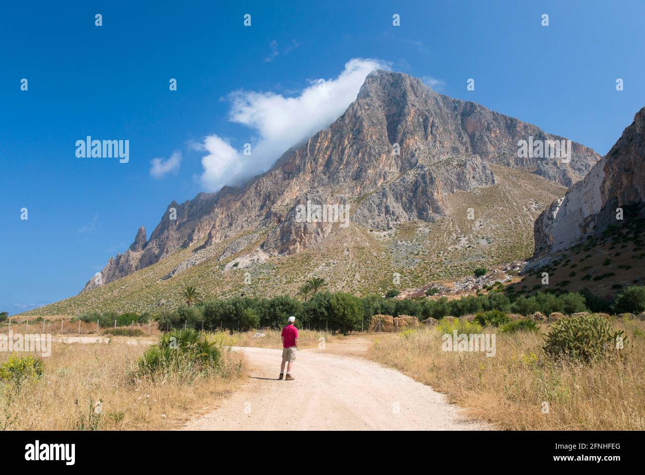 Custonaci, Trapani, Sicily, Italy. Solitary visitor on dusty track between fields admiring view of the towering south face of Monte Cofano. Stock Photo