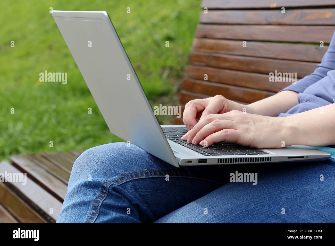 Woman sitting with a laptop on her lap on a wooden bench in park. Female hands on keyboard close up, concept of remote work outdoors, freelancer Stock Photo
