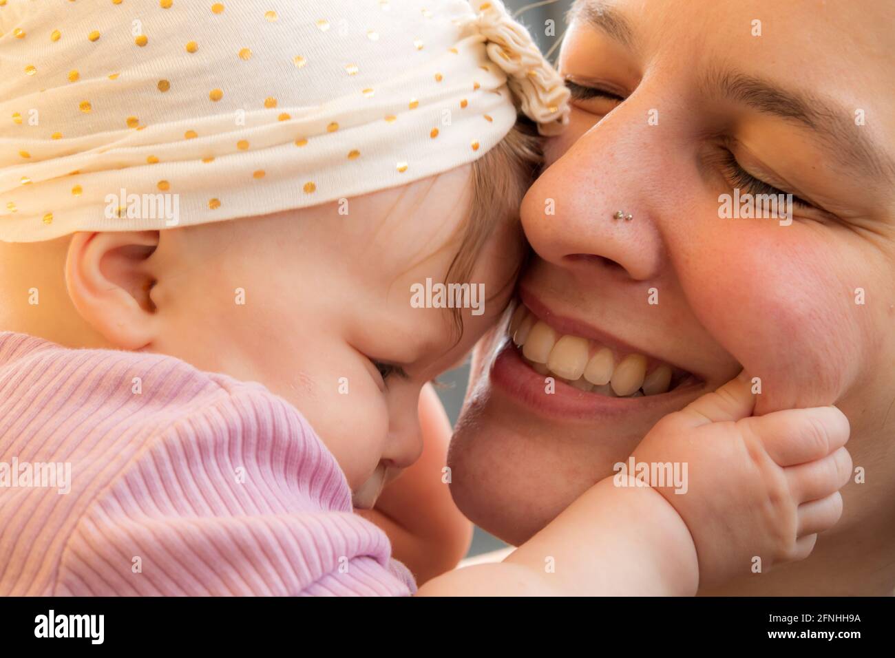 Close up of young mother with 5 month old baby, hugging, smiling, loving expression.  Baby with hat, holding mother's cheek. Stock Photo