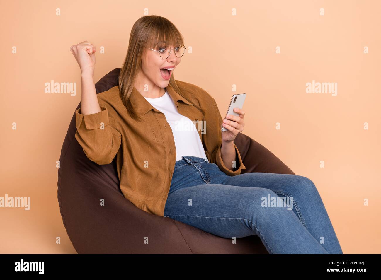 Photo Of Amazed Victorious Young Woman Look Phone Raise Fist Sit Bean Bag Isolated On Beige Color Background Stock Photo Alamy