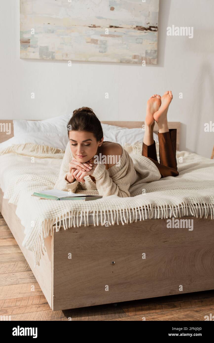 barefoot young woman reading book while resting on bed Stock Photo