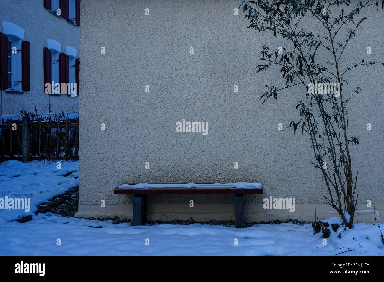 A snow covered bench in front of a house wall on which the branches of a plant grow up. Stock Photo