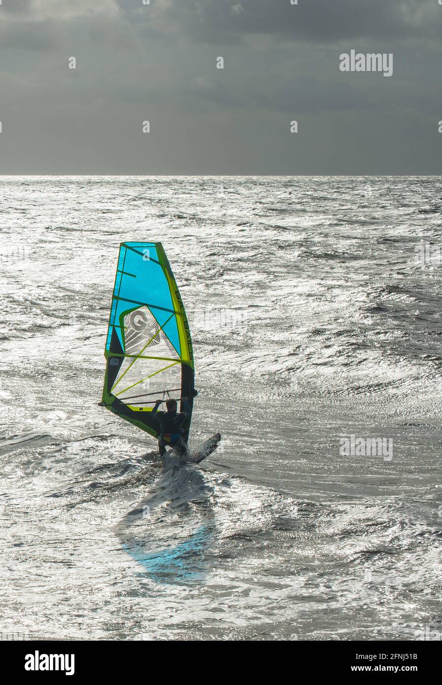 Striking image of a single windsurfer with blue sail speeding along with a dark sky background set against a silvery sea Stock Photo