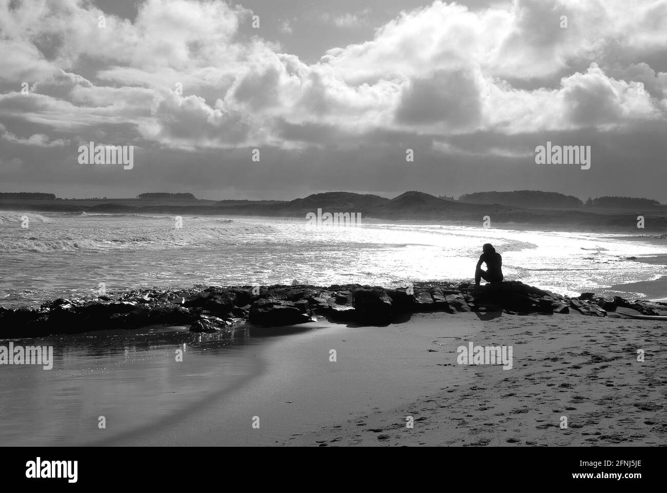 A mono image of a single figure in silhouette sitting on rocks at the coast and looking out to sea with dunes and drifting clouds as background Stock Photo