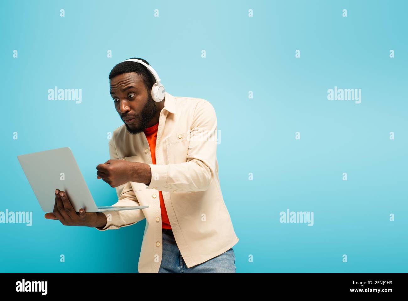 Angry African American Man In Headphones Showing Clenched Fist Near Laptop On Blue Background