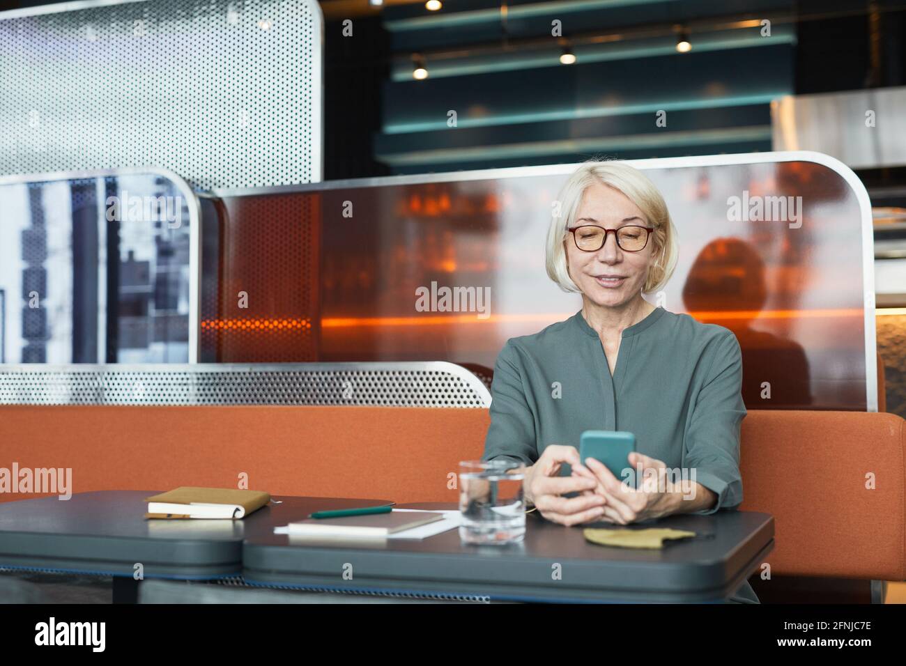 Smiling Caucasian blond-haired senior woman in eyeglasses sitting at table with organizer and checking phone in modern cafe Stock Photo