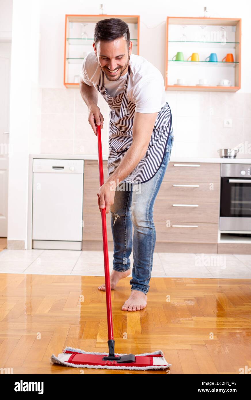 Handsome guy mopping parquet floor in front of kitchen cabinets and countertop at home Stock Photo