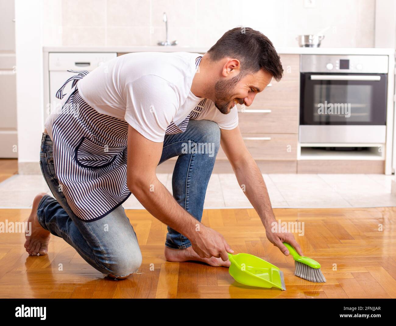 Handsome man sweeping floor with dust pan in front of kitchen cabinets at home Stock Photo